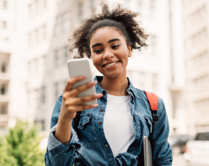 Una alegre estudiante afroamericana usa su teléfono móvil para enviar mensajes de texto y aprender en una aplicación educativa, parada afuera cerca de un edificio universitario. Nuevo concepto de aplicación para tu celular.